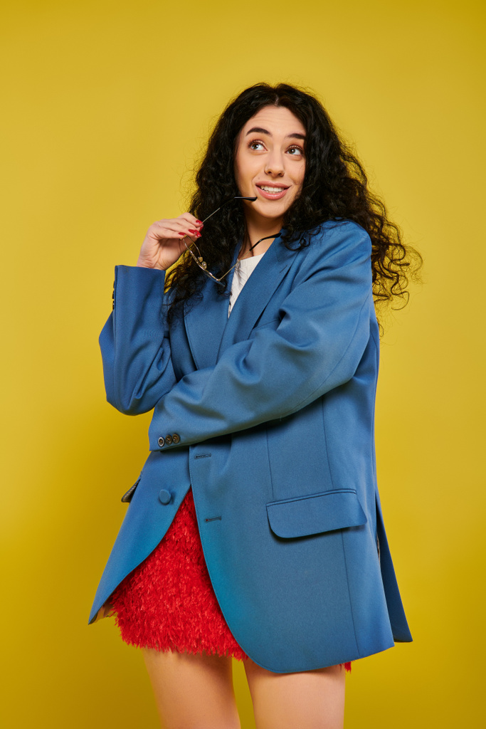 Una joven morena con el pelo rizado posa en una elegante chaqueta azul y falda roja, expresando emociones sobre un fondo amarillo. - Foto, Imagen