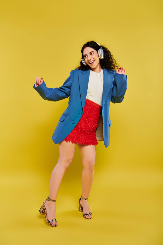 Brunette vrouw met krullend haar poseert in stijlvolle blauwe jas en rode rok, uitstralen vertrouwen en elegantie tegen een gele achtergrond. - Foto, afbeelding