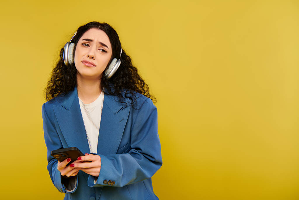 Una mujer morena con el pelo rizado usando auriculares y sosteniendo un teléfono celular, perdido en la música contra un fondo amarillo. - Foto, imagen