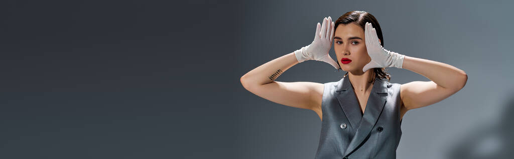 Стильна молода жінка позує в елегантному костюмі з жилетом, руками на голові, висловлюючи емоції в студії на сірому фоні. - Фото, зображення
