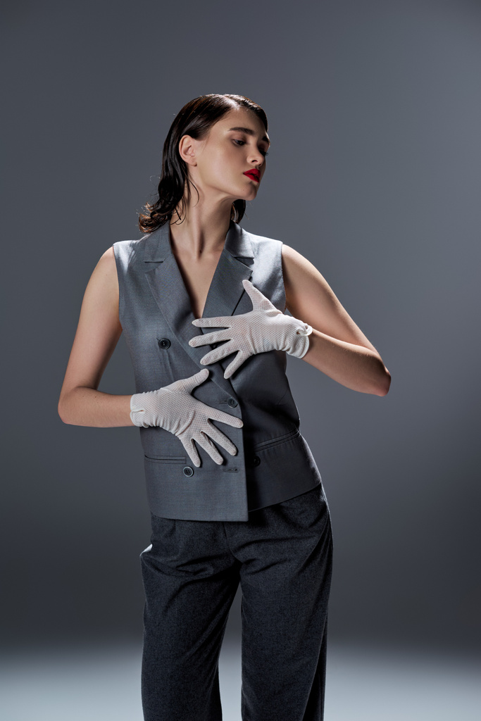 Elegante giovane donna colpisce una posa in un elegante abito grigio con un gilet, completato da guanti bianchi, in uno studio sullo sfondo grigio. - Foto, immagini