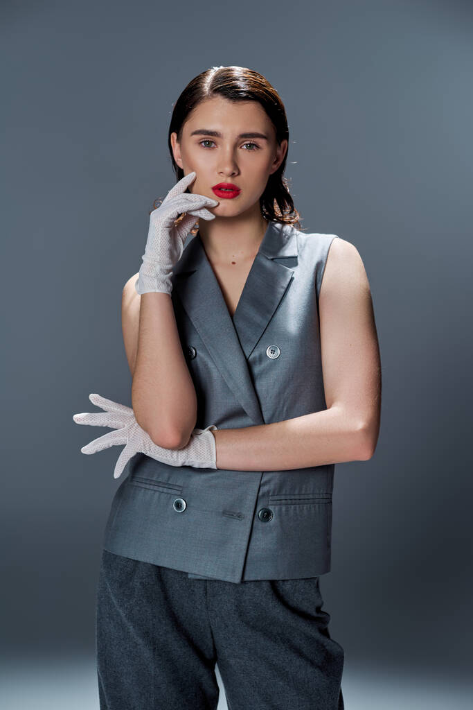 Een stijlvolle jonge vrouw poseert in een elegant grijs pak en witte handschoenen tegen een studio achtergrond. - Foto, afbeelding