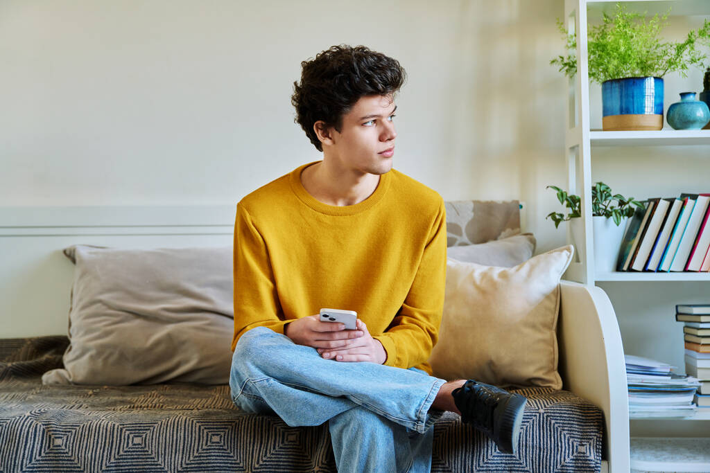 Young guy holding smartphone in hands sitting on couch at home. Handsome university college student 19-20 years old in yellow with curly hair. Technologies, mobile apps, study communication leisure - Photo, Image