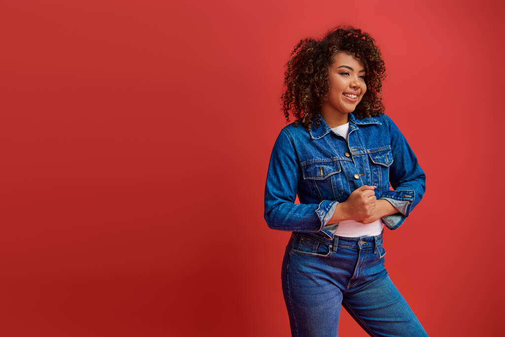 joyous elegant african american woman in stylish denim outfit looking away on red backdrop - Photo, Image