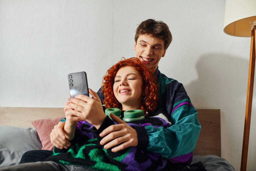 good looking joyful man with headphones looking at mobile phone with his cheerful girlfriend on bed - Photo, Image
