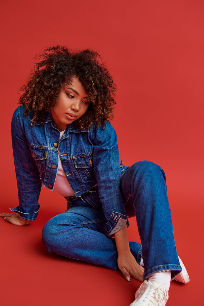 elegant young african american woman in stylish denim outfit looking away on red vibrant backdrop - Photo, Image
