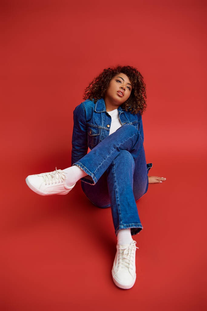заманчивая молодая африканская американка в стильном джинсовом наряде, смотрящая на красный фон - Фото, изображение