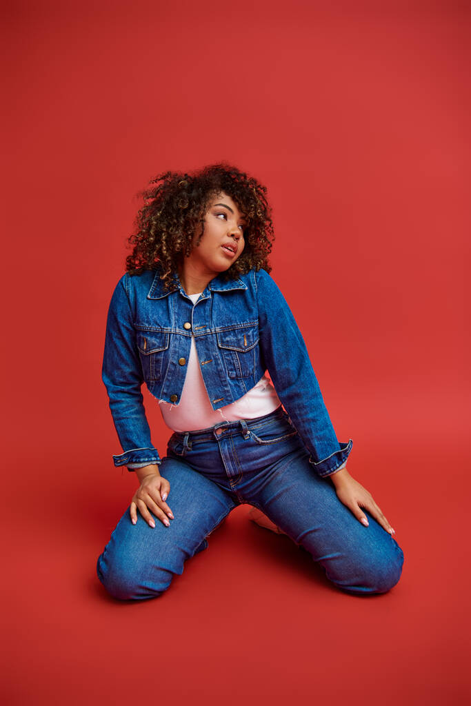 приваблива молода афроамериканська жінка в стильному джинсовому вбранні, дивлячись далеко на червоний фон - Фото, зображення