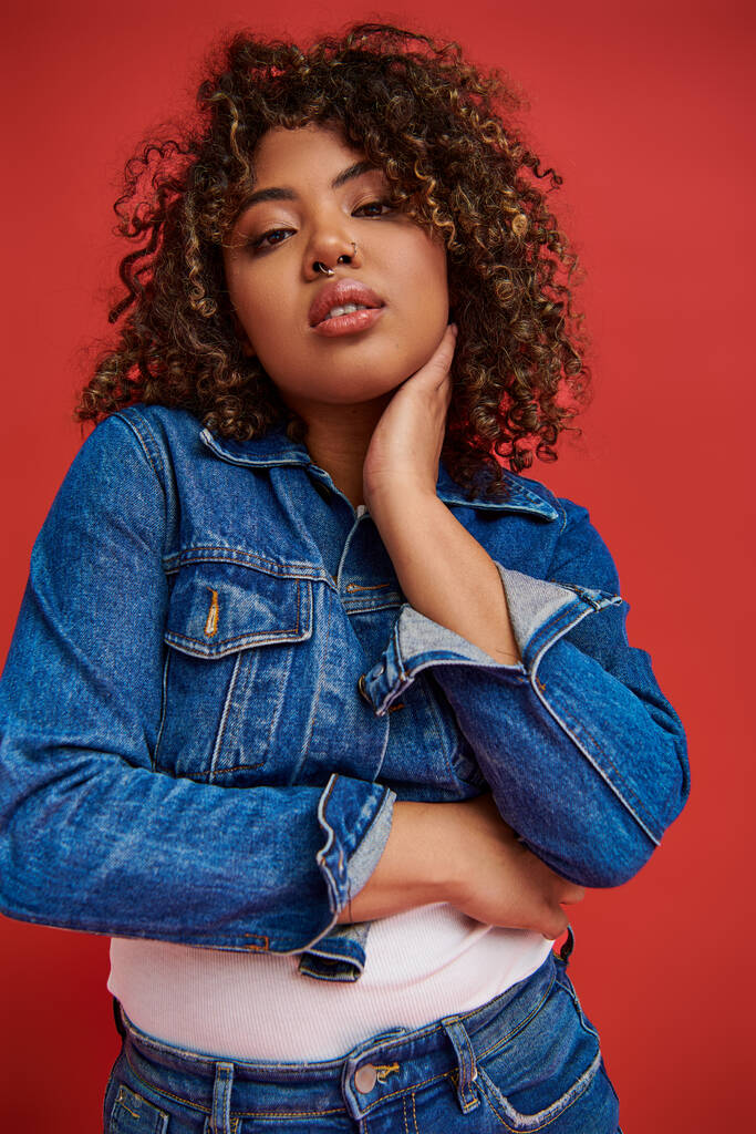 attractive african american woman in stylish denim outfit looking at camera on vibrant red backdrop - Photo, Image