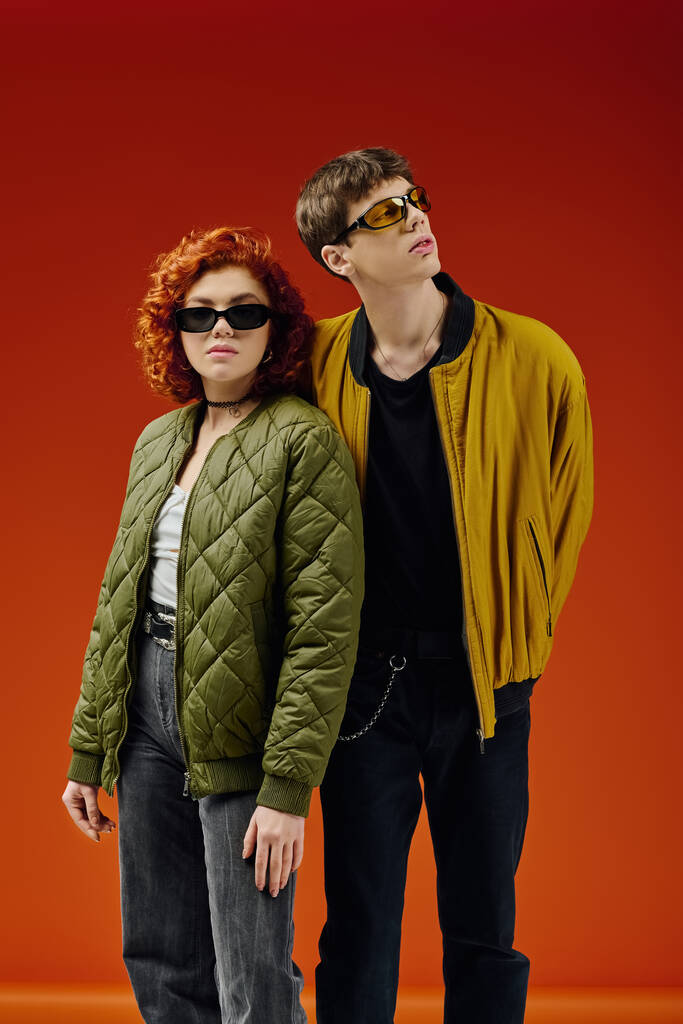 стильная привлекательная пара в модных нарядах с солнцезащитными очками, позирующими вместе на красном фоне - Фото, изображение