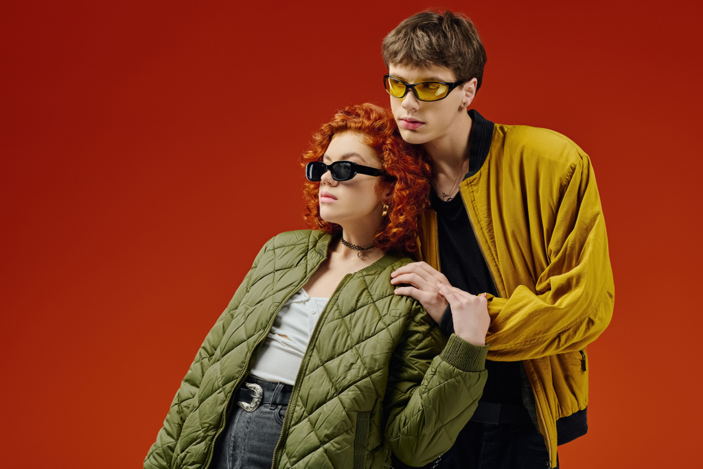 atractiva pareja de moda con gafas de sol frescas en trajes cómodos posando sobre fondo rojo juntos - Foto, Imagen