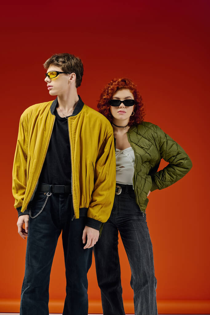 attractive stylish couple with cool sunglasses in cozy attires posing on red backdrop together - Photo, Image