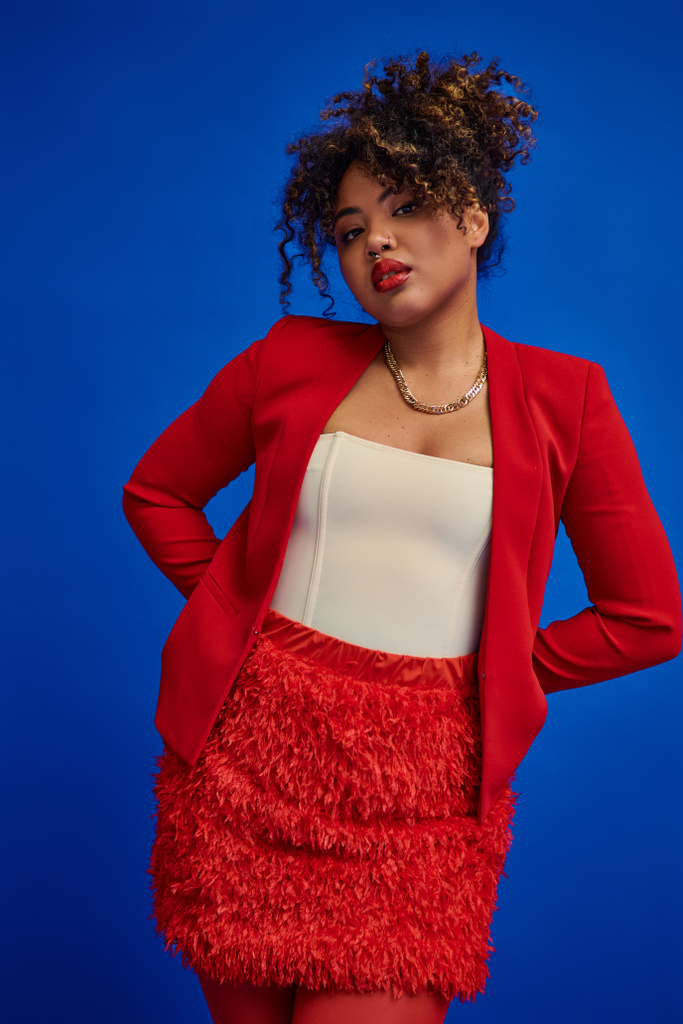dapper young african american woman in red blazer with curly hair looking at camera, blue backdrop - Photo, Image