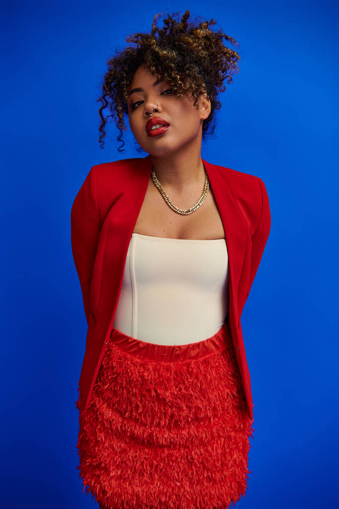 exquisite chic african american woman in red blazer with curly hair looking at camera, blue backdrop - Photo, Image