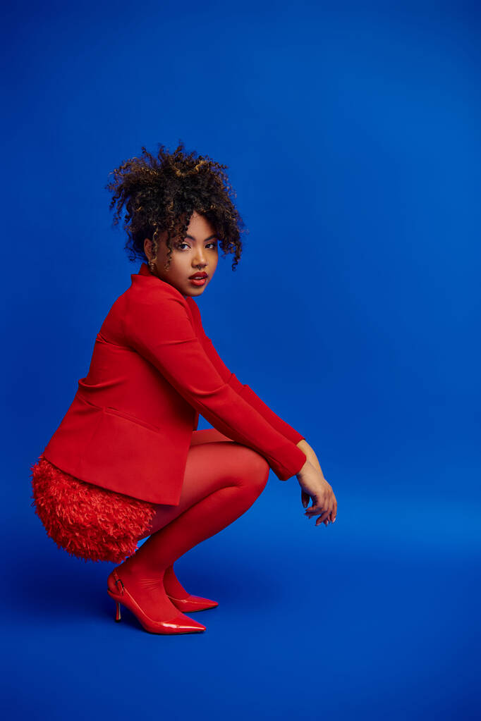 flancos elegáns afro-amerikai nő piros blézer göndör haj néz kamera, kék háttér - Fotó, kép