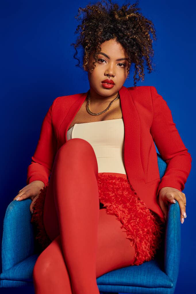 encantadora mujer afroamericana elegante en traje rojo inteligente sentado en la silla azul y mirando a la cámara - Foto, imagen