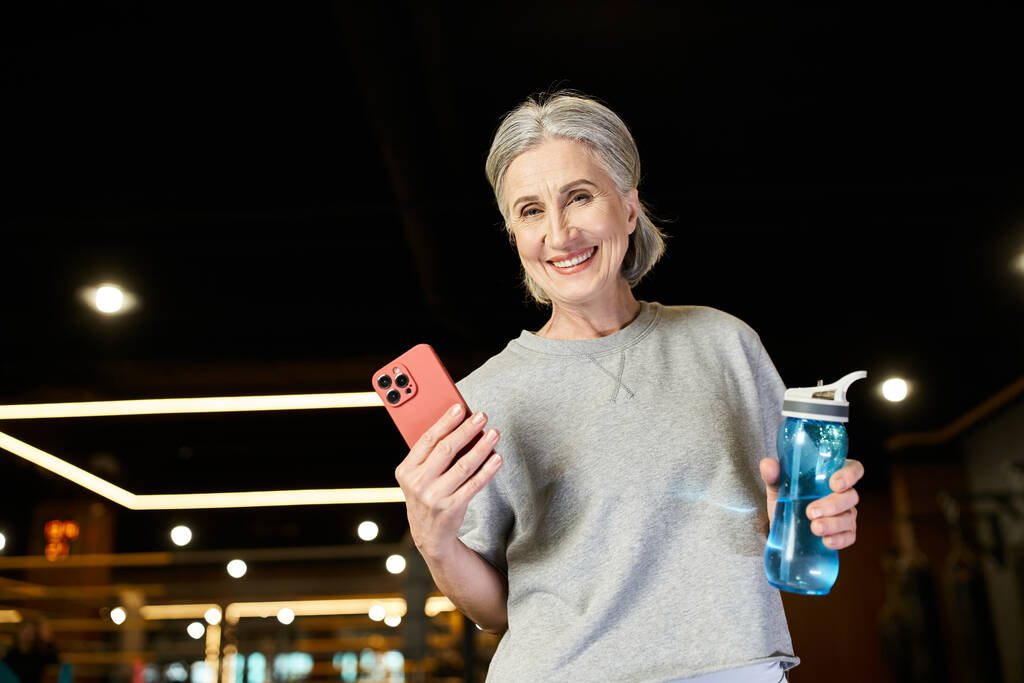 spokojený starší sportovkyně s šedými vlasy drží láhev vody a telefon a usmívá se na kameru - Fotografie, Obrázek
