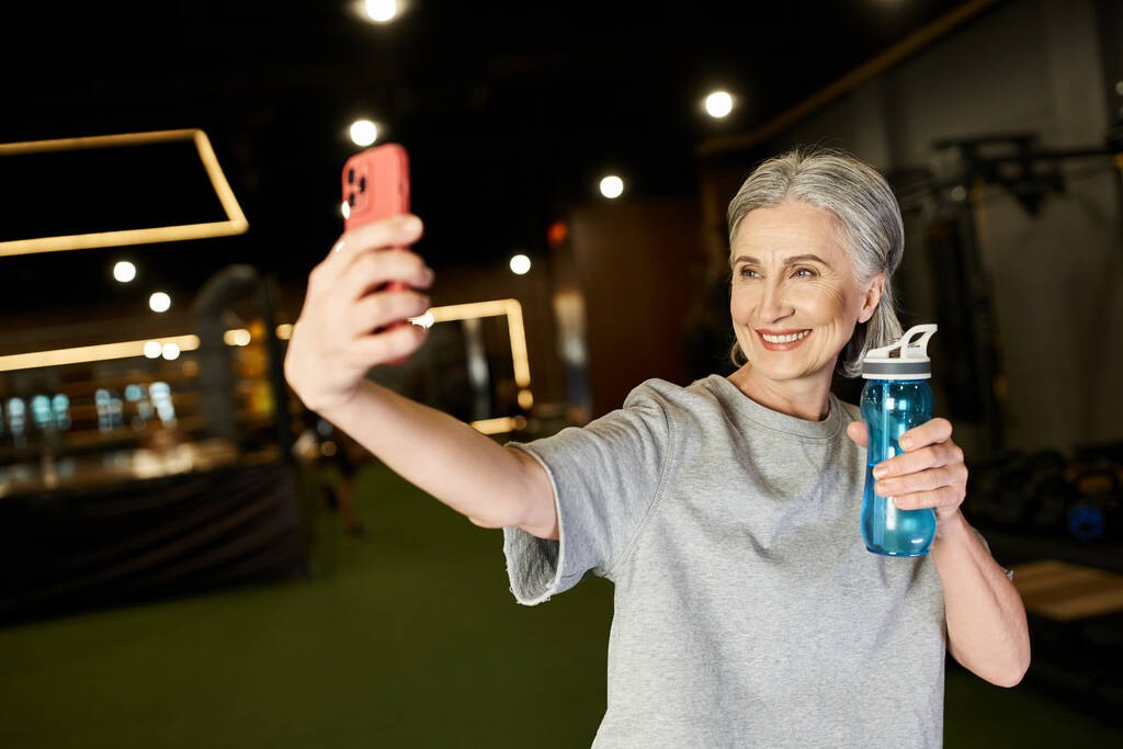 vesele dobře vypadající senior sportovkyně v útulném oblečení přičemž selfie v tělocvičně a držení láhve s vodou - Fotografie, Obrázek