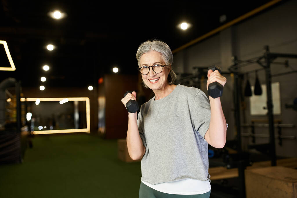 веселая старшая спортсменка с седыми волосами, тренирующаяся с гантелями и улыбающаяся в камеру - Фото, изображение