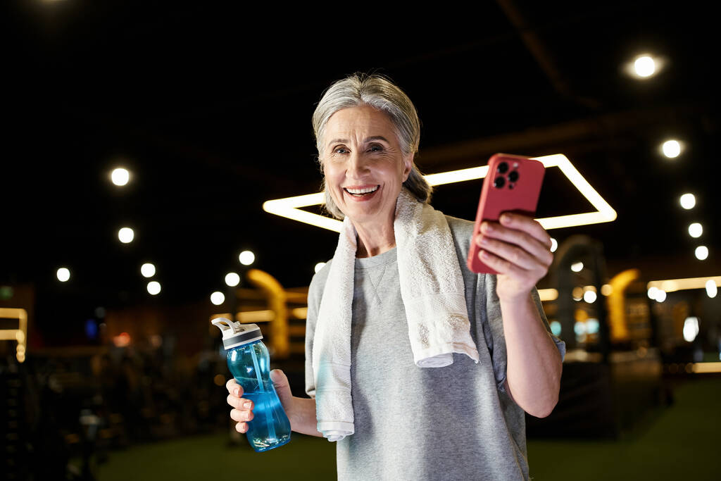 radosna starsza kobieta z ręcznikiem na ramionach trzymająca telefon i butelkę i uśmiechająca się do kamery - Zdjęcie, obraz