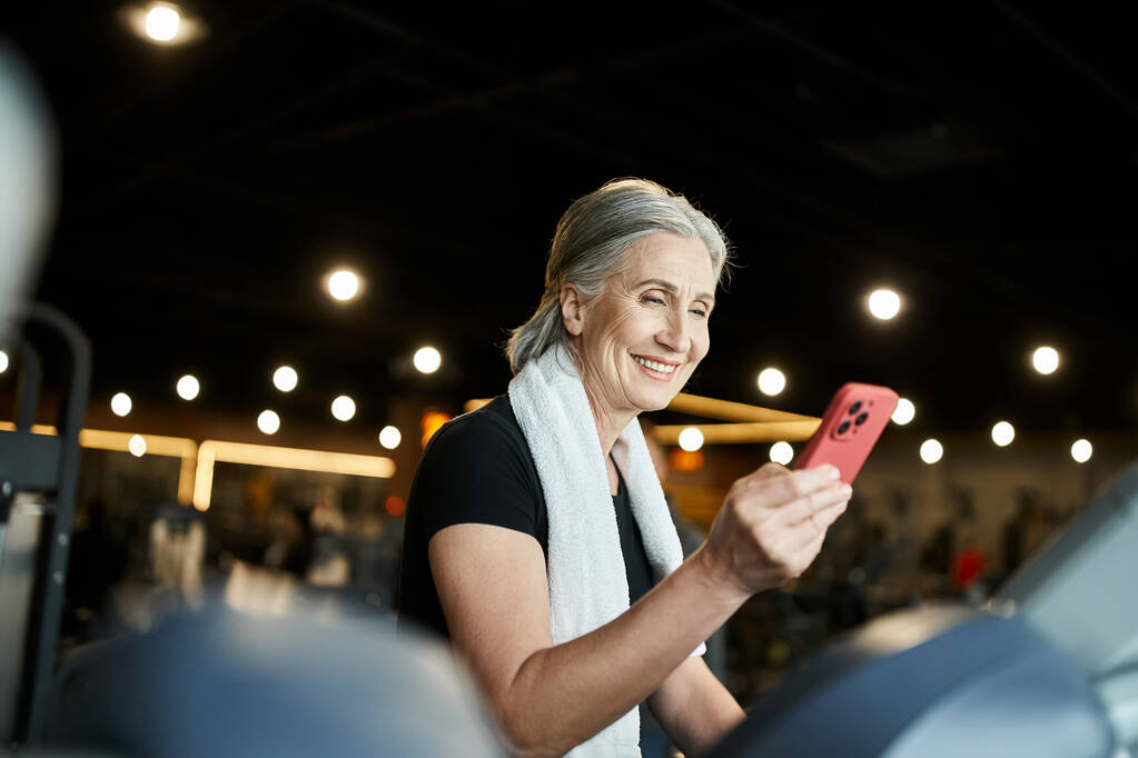 vrolijke volwassen sportvrouw met grijs haar het hebben van video oproep tijdens de training op loopband in de sportschool - Foto, afbeelding