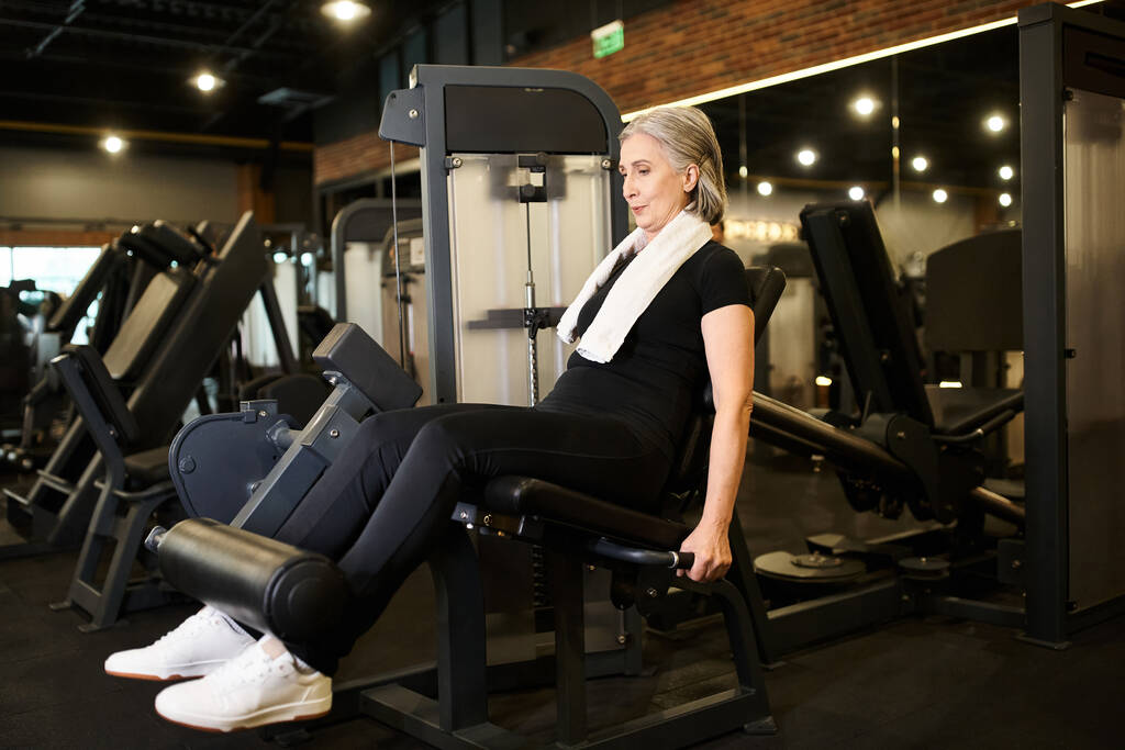 athlétique mature femme aux cheveux gris dans l'entraînement confortable de vêtements de sport sur la machine à boucler les jambes tout en gymnase - Photo, image