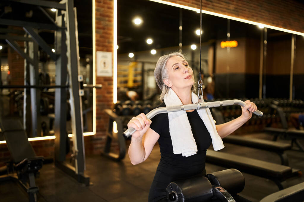 atraktivní sportovní starší žena v útulném oblečení cvičení aktivně s lats pulldown stroj v tělocvičně - Fotografie, Obrázek