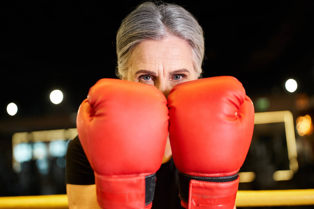 Spor kıyafetleri içinde oldukça neşeli olgun bir kadın boks eldivenleriyle ringde antrenman yapıyor ve kameraya gülümsüyor. - Fotoğraf, Görsel