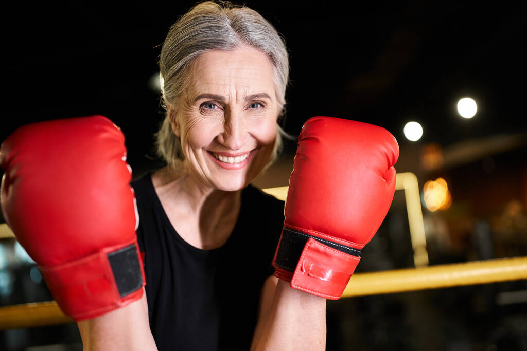 привлекательная веселая пожилая женщина в спортивной одежде в боксёрских перчатках, тренирующаяся на ринге и улыбающаяся в камеру - Фото, изображение