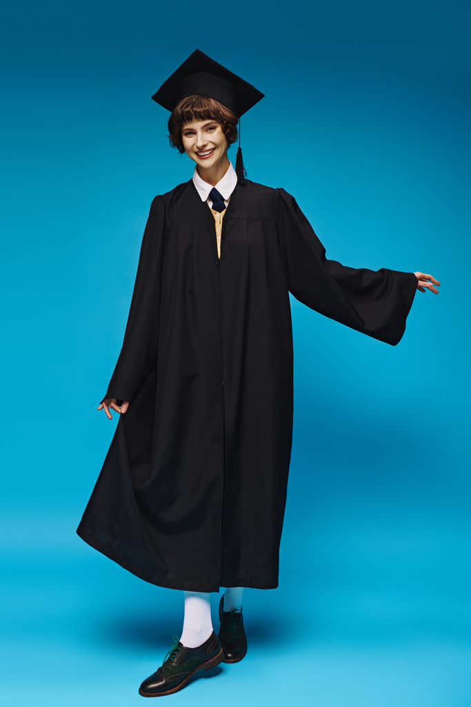 concepto de graduación, chica universitaria feliz en gorra académica y vestido de pie sobre fondo azul - Foto, imagen