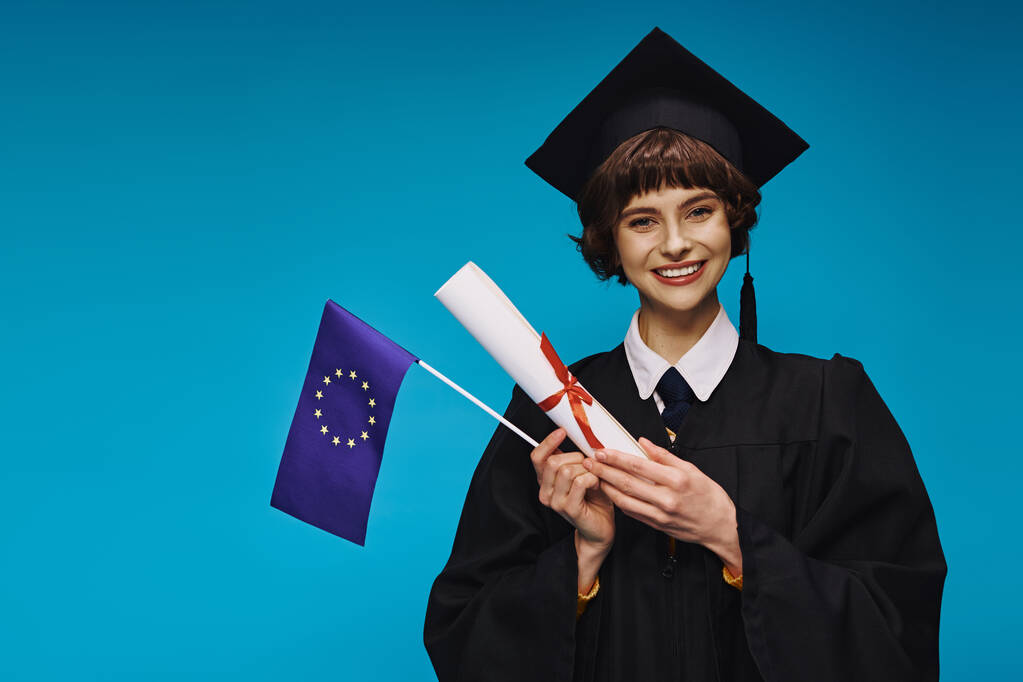 vrolijk afgestudeerd college meisje in toga en academische pet met diploma en EU-vlag op blauwe achtergrond - Foto, afbeelding