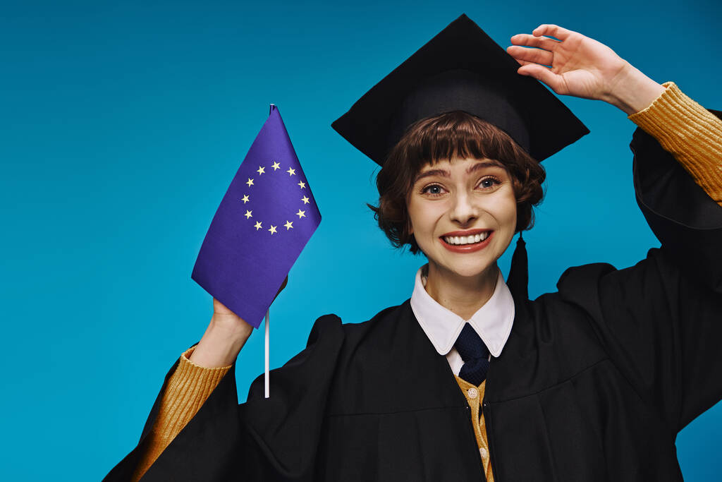 chica graduada en vestido y gorra sosteniendo la bandera de la UE y sonriendo en el telón de fondo azul, educación europea - Foto, Imagen