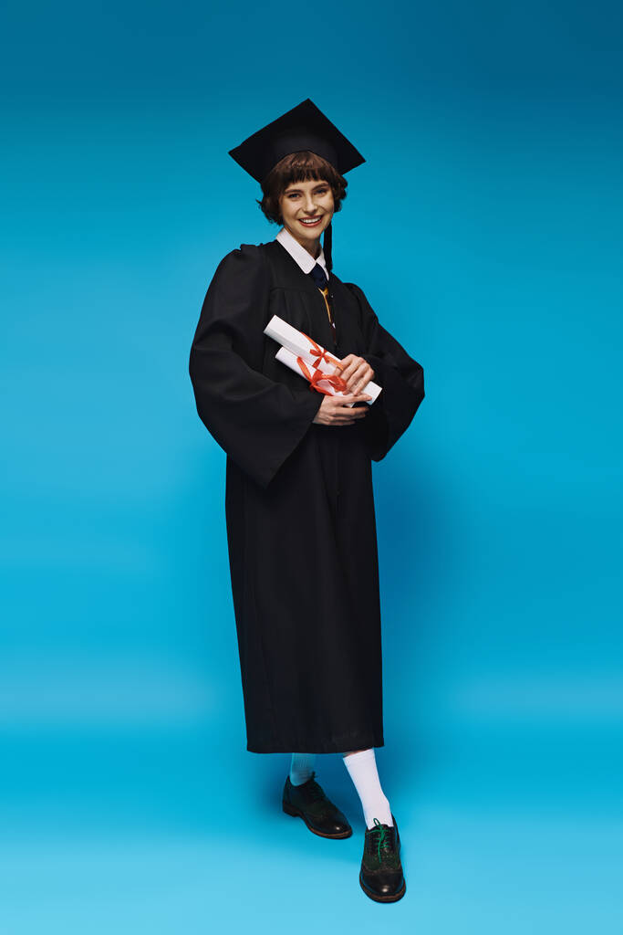 happy grad college girl in gown and academic cap holding diplomas with pride, blue background - Photo, Image