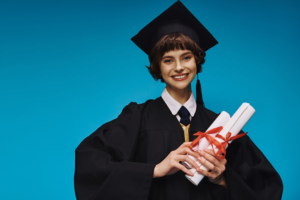feliz graduado chica universitaria en vestido y la gorra académica que sostiene diplomas con orgullo en azul - Foto, Imagen