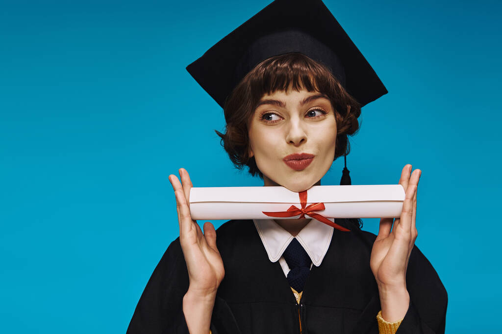 gelukkig grad college meisje in toga en academische pet pruilen lippen en het houden van haar diploma met trots - Foto, afbeelding