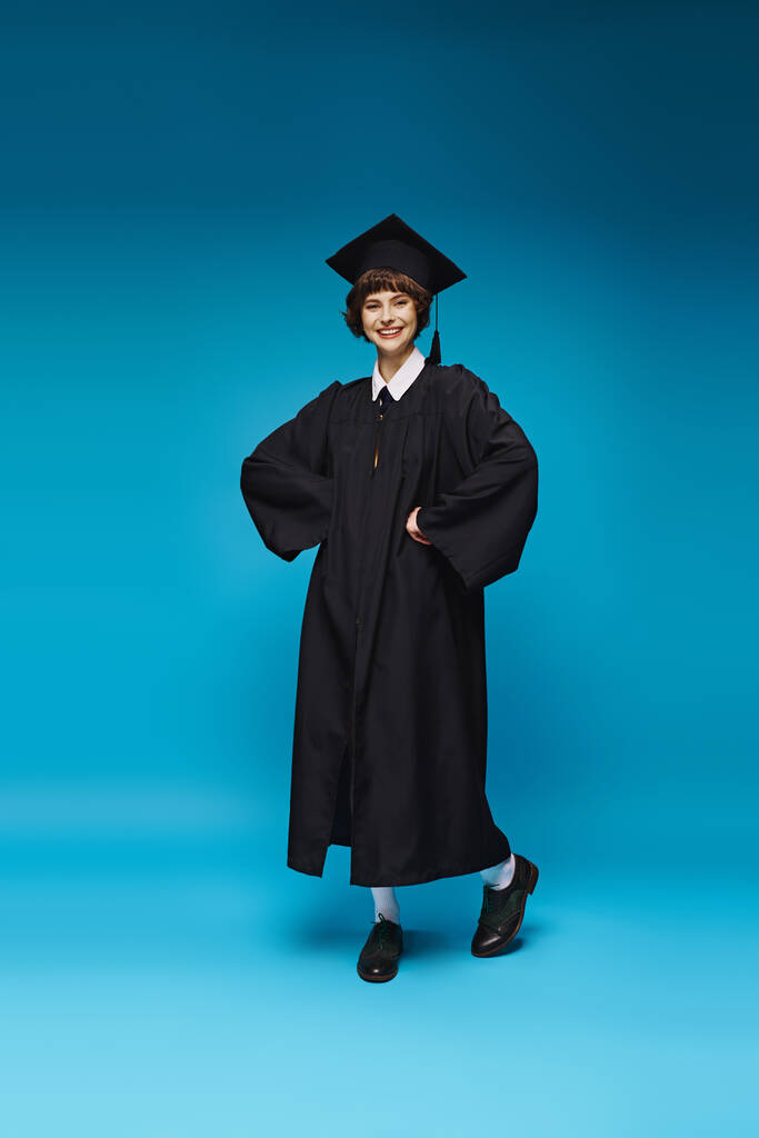 alegre graduado menina da faculdade no vestido e boné sorrindo no fundo azul, realização - Foto, Imagem
