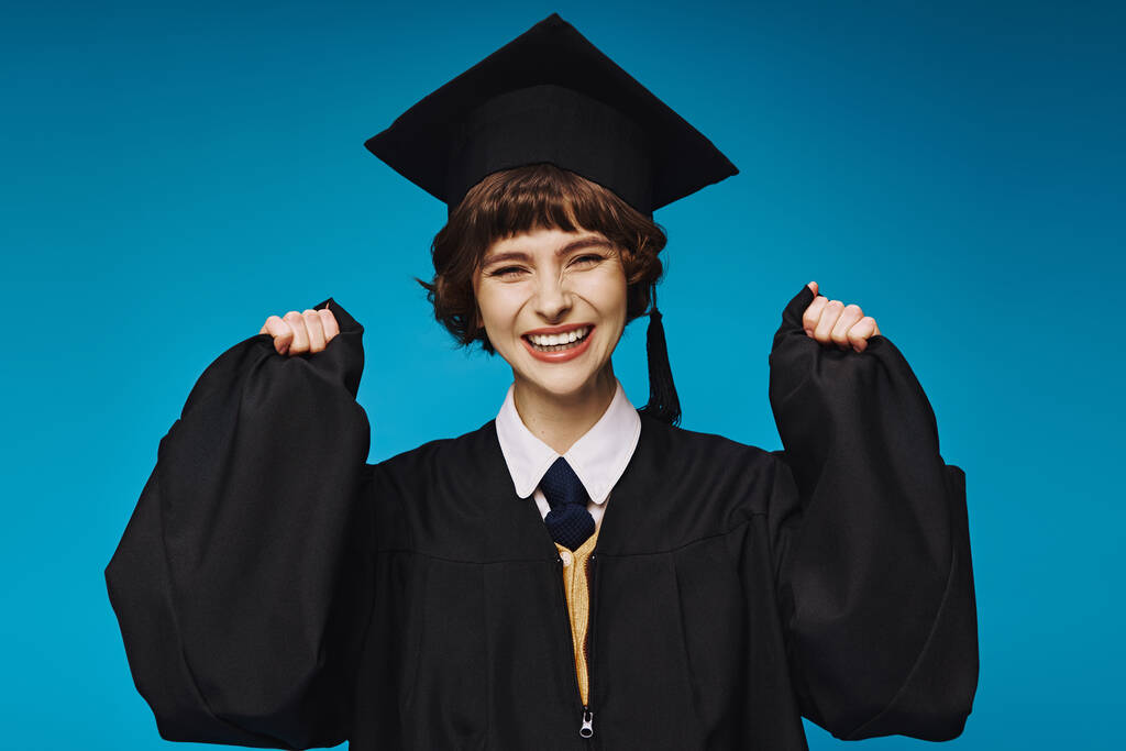 jeune brunette diplômée fille de l'université en chapeau académique se sentant heureux sur fond bleu, succès - Photo, image
