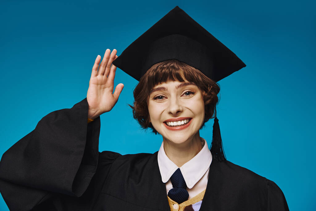 Akademik şapkalı üniversiteli genç ve neşeli kız el sallıyor, mavi arkaplan. - Fotoğraf, Görsel