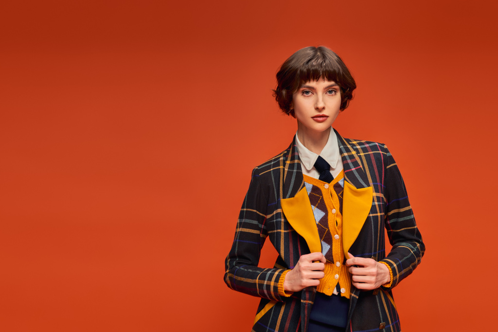 stylish student with short hair posing in checkered blazer on orange background, college uniform - Photo, Image