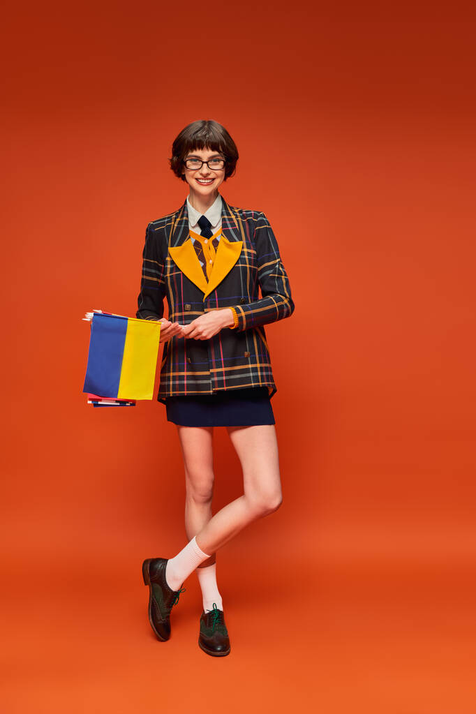 happy young college girl in uniform and glasses holding Ukrainian flag on orange background - Photo, Image