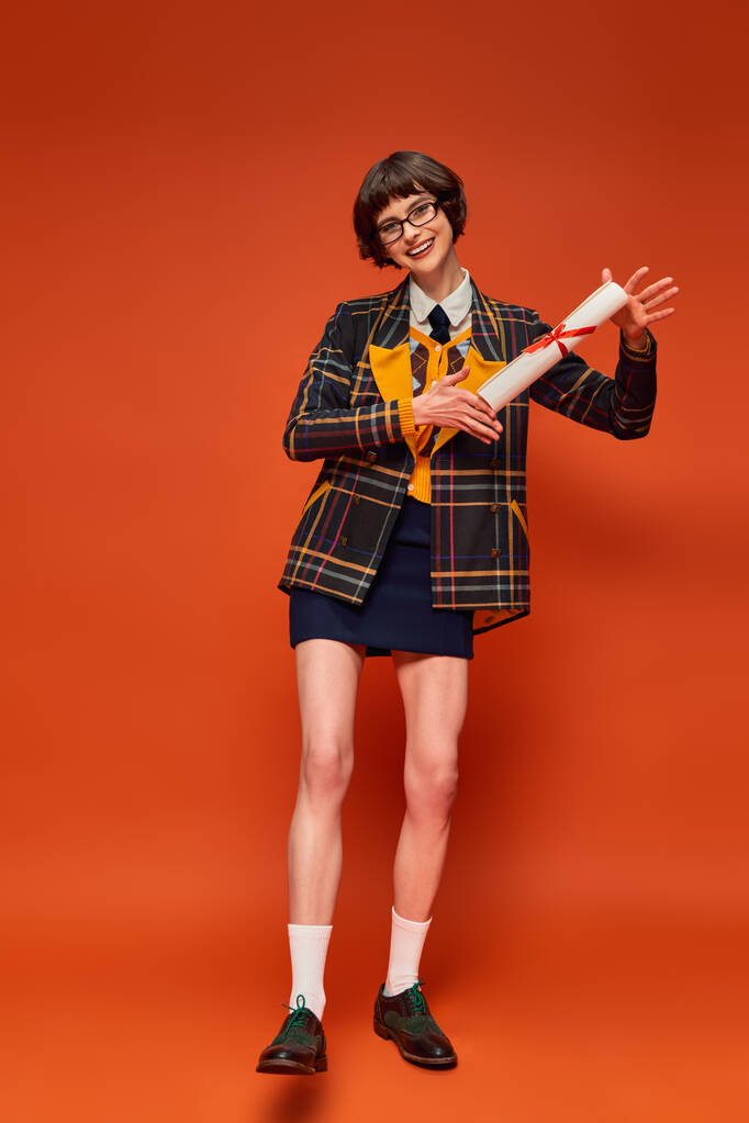 excitada chica universitaria graduada en uniforme y gafas sosteniendo su diploma en vibrante fondo naranja - Foto, imagen
