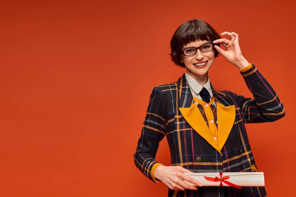 fröhliches Hochschulmädchen in Uniform und Brille mit Diplom vor leuchtend orangefarbenem Hintergrund - Foto, Bild