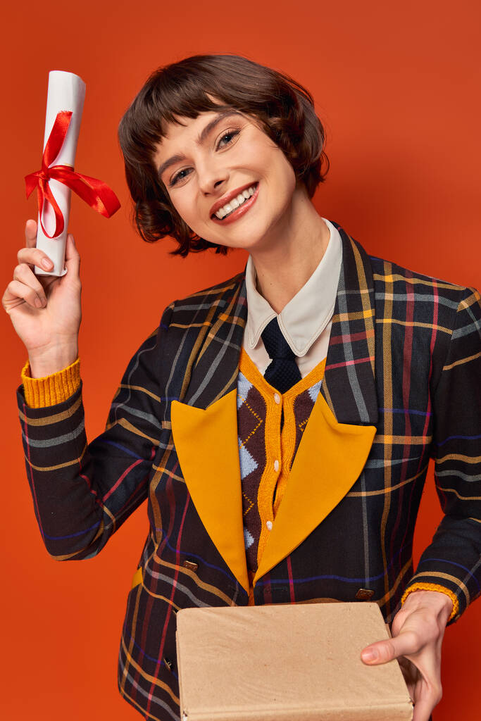 portrait of excited college girl in checkered uniform holding books and diploma on orange backdrop - Photo, Image