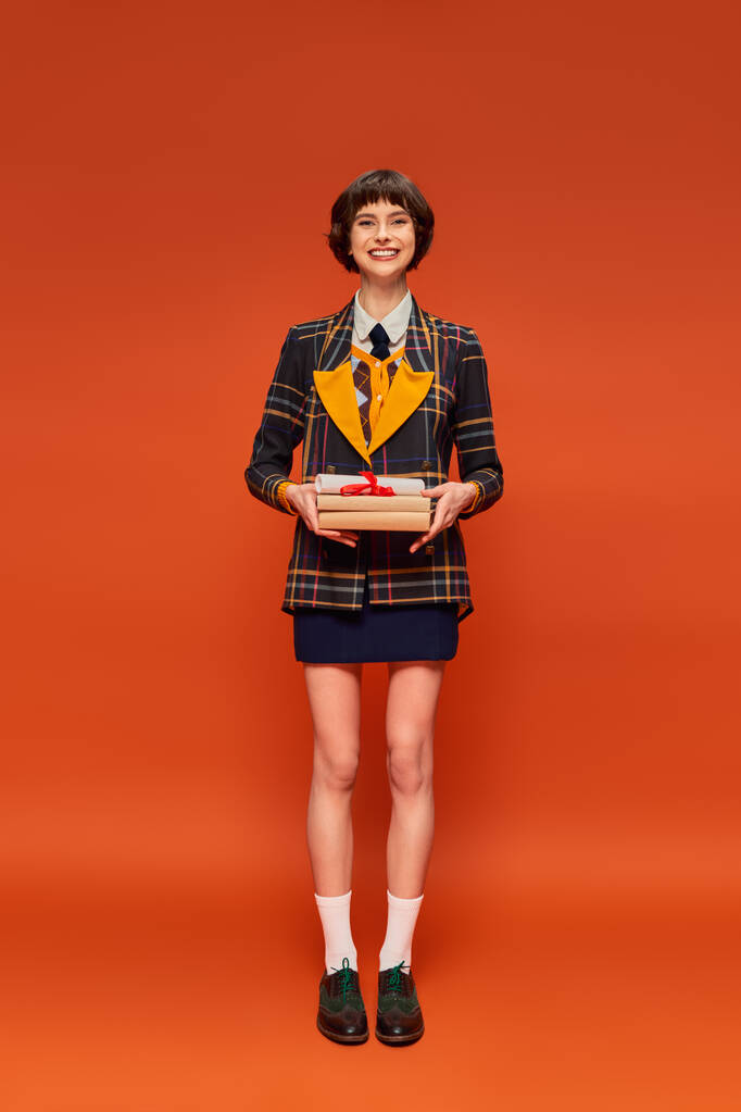 longitud completa de estudiante positivo en uniforme de la universidad de pie con libros sobre fondo naranja - Foto, Imagen