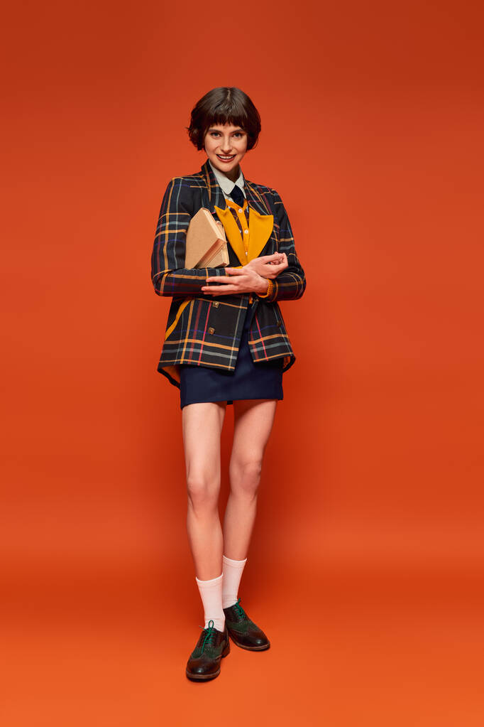 portrét pozitivního studenta v univerzitní uniformě stojící s knihami na oranžovém pozadí, znalosti - Fotografie, Obrázek
