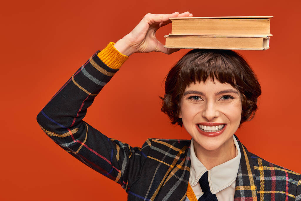 positive college girl in uniform holding stack of books on hand on orange background, knowledge - Photo, Image