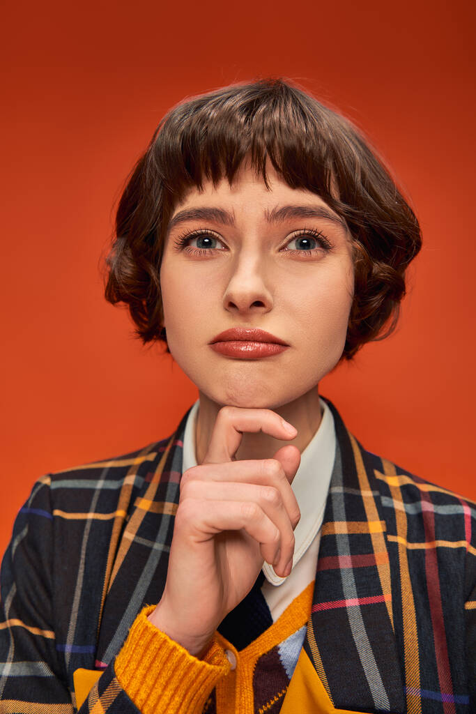 riflessivo college girl con i capelli corti posa in uniforme a scacchi su sfondo arancione - Foto, immagini
