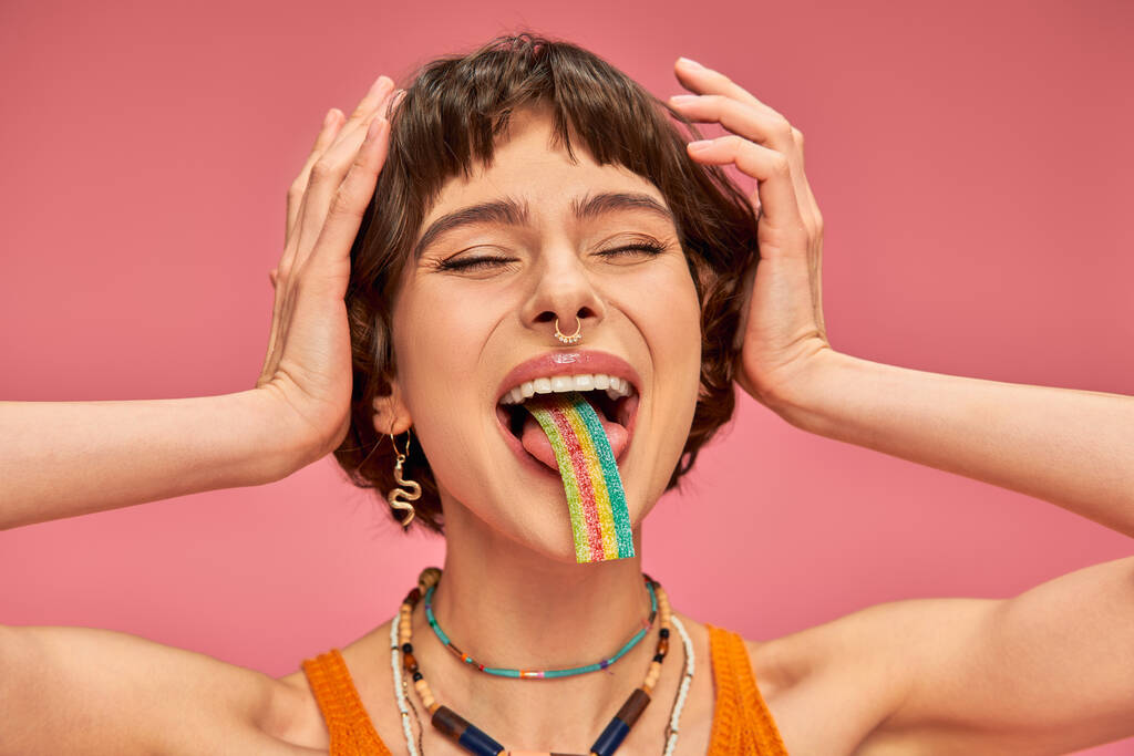 excité jeune femme dans sa vingtaine dégustation sucré et aigre bande de bonbons sur sa langue, fond rose - Photo, image