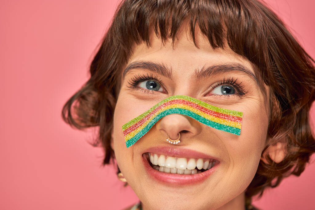 счастливая женщина в возрасте 20 лет со сладкими и кислыми конфеты полоску на носу на розовом фоне - Фото, изображение