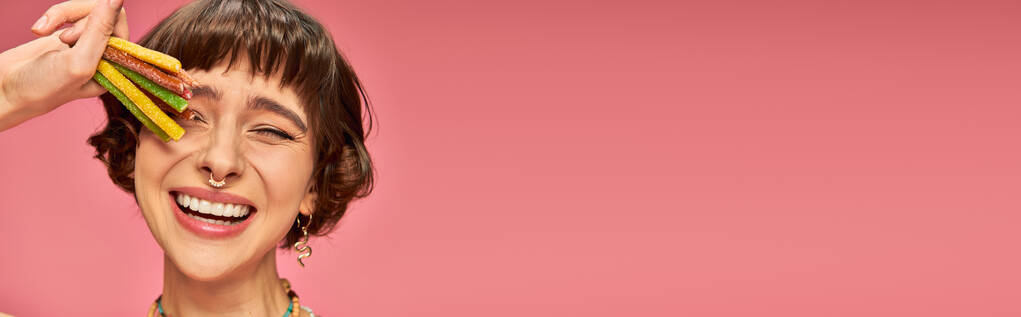 весела молода жінка в 20 років посміхається і тримає солодкі і кислі цукерки в руці на рожевому, банер - Фото, зображення